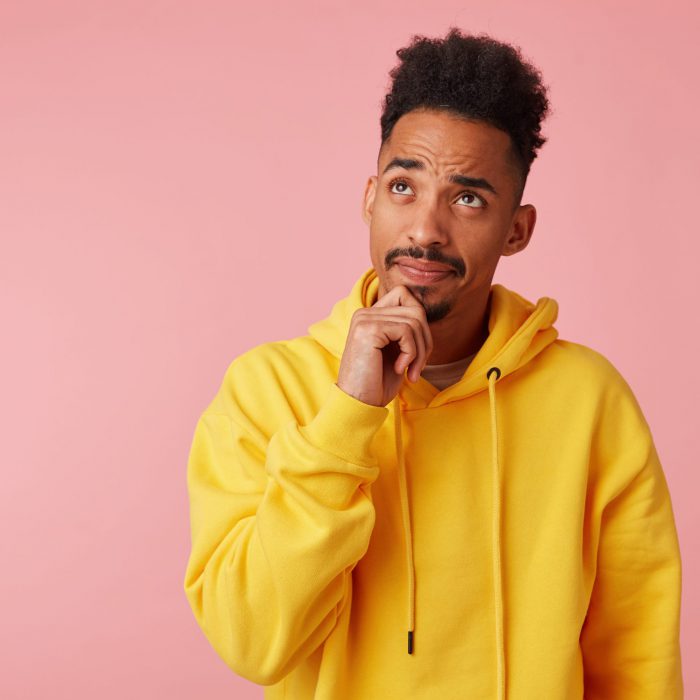 Young thinking african american guy in yellow hoodie, looks up and touches chin, stands over pink background with copy space.